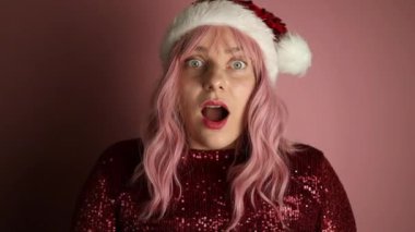Noel Baba şapkası takan genç bir kadın avuç içi açık, çığlık çığlığa evet diyor ve açık pembe renkli arka planda izole edilmiş zaferi kutluyor. Yüksek kaliteli FullHD görüntüler