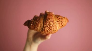 Pembe arka planda taze ve lezzetli pişmiş altın kruvasan tutan kadın eli. Yüksek kaliteli FullHD görüntüler
