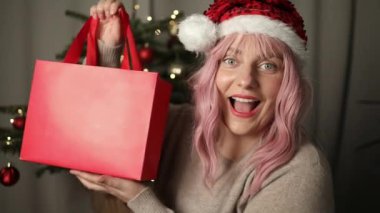 Genç pembe saçlı kadın evde Noel kutlamalarında kendine güvenen bir şekilde Noel hediyesini tutuyor. Yüksek kaliteli FullHD görüntüler