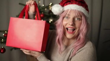 Genç pembe saçlı kadın evde Noel kutlamalarında kendine güvenen bir şekilde Noel hediyesini tutuyor. Yüksek kaliteli FullHD görüntüler