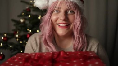 Neşeli, genç ve güzel bir kadın. Elleri rahat bir süveter içinde. Üzerinde yılbaşı hediyesi, arka planda ışıklarla Noel ağacı resmi var. Kurdeleli zarif bir kadın. 