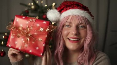 Neşeli, genç ve güzel bir kadın rahat bir süveter içinde yılbaşı hediyesini açıyor ve arka planda ışıkları olan bir noel ağacı var. Kurdeleli zarif bir kadın. 