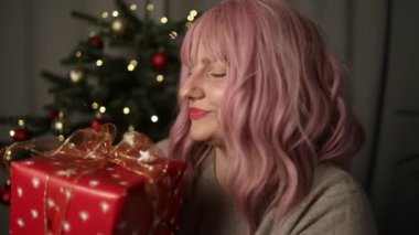 Neşeli, genç ve güzel bir kadın. Elleri rahat bir süveter içinde. Üzerinde yılbaşı hediyesi, arka planda ışıklarla Noel ağacı resmi var. Kurdeleli zarif bir kadın. 