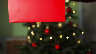 Kameraya kırmızı hediyesini gösteren mutlu Asyalı kadın. Noel süsleriyle süslü bir ağacın yanında oturan gülümseyen kadın, Noel arifesi, yeni yıl arifesi. Yüksek kalite FullHD