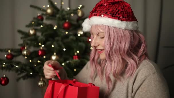 エキサイティングな幸せな美しいサンタ帽子ピンクの髪の少女は 注文や贈り物で赤いプレゼントを開き 驚いた若い女性は家族からプレゼントを得ました エクスプレスデリバリーサービス ライフイベントコンセプト — ストック動画