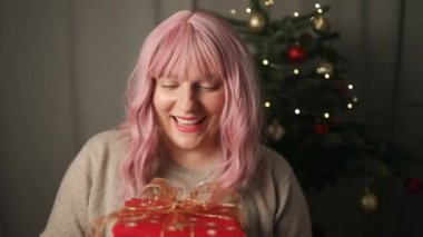 Neşeli, genç ve güzel bir kadın rahat bir süveter içinde yılbaşı hediyesini açıyor ve arka planda ışıkları olan bir noel ağacı var. Kurdeleli zarif bir kadın. 