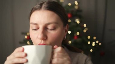 Güzel genç bir kadın Noel arifesinde evdeki süslü Noel ağacının yanında kanepede otururken kahve içiyor. Yüksek kaliteli FullHD görüntüler
