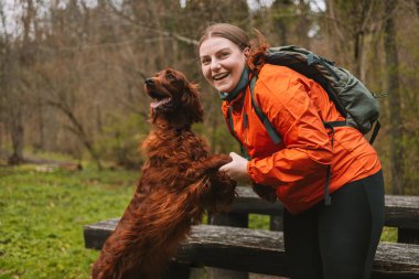 Sırt çantalı mutlu genç bir kadın köpekle seyahat ediyor. Genç bir dişi sevgili köpeğine sarılıyor. Kız ve İrlanda setter bahar ormanında gün batımında. Yüksek kalite fotoğraf