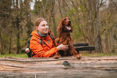 Sırt çantalı mutlu genç bir kadın köpekle seyahat ediyor. Genç bir dişi sevgili köpeğine sarılıyor. Kız ve İrlanda setter bahar ormanında gün batımında. Yüksek kalite fotoğraf