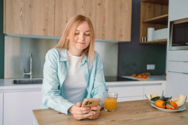 Evdeki mutfakta bir bardak su içerken orta yaşlı, gülümseyen bir kadının fotoğrafı.