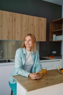 Evdeki mutfakta bir bardak su içerken orta yaşlı, gülümseyen bir kadının fotoğrafı.