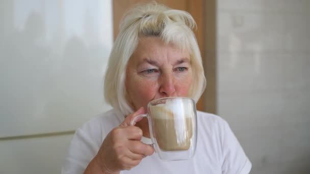 キッチンでコーヒーラテを飲む窓に座っている白人60代の女性の近く カフェでコーヒーを飲むことを楽しむ美しい女性 — ストック動画