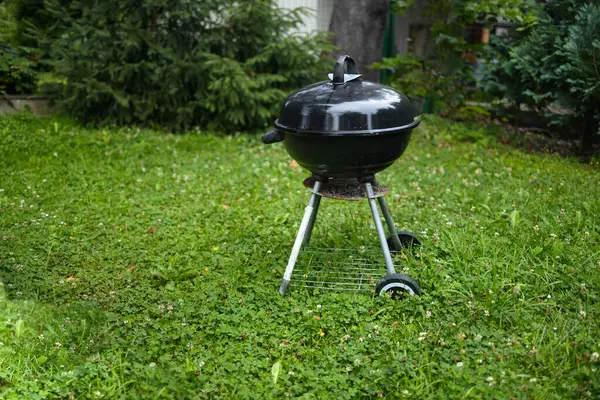 Side view of black barbecue grill with cover in front of house or backyard and green lawn. Portable barbecue grill for outdoor cooking.