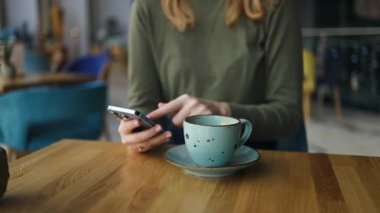 Kahve dükkanında kahve masasında otururken elinde akıllı telefon tutan ve cep telefonunu kaydıran bir kadının görüntüsü. Çalışan insanlar pazarlama işiyle, teknolojiyle iletişime geçiyor. Yüksek kalite FullHD