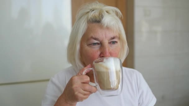 キッチンでコーヒーラテを飲む窓に座っている白人60代の女性の近く カフェでコーヒーを飲むことを楽しむ美しい女性 — ストック動画