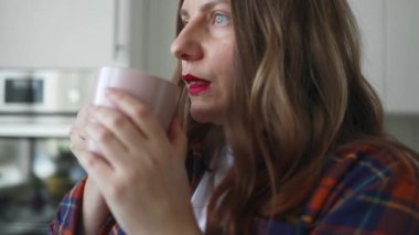 Güneşli bir sabahta pencerenin yanında panjurlu sıcak kahve ya da çay içen genç bir kadın. Kadın evde dinleniyor, sıcak bir şeyler içiyor ve şehir manzarasına bakıyor. Yüksek kaliteli FullHD görüntüler