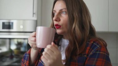 Güneşli bir sabahta pencerenin yanında panjurlu sıcak kahve ya da çay içen genç bir kadın. Kadın evde dinleniyor, sıcak bir şeyler içiyor ve şehir manzarasına bakıyor. Yüksek kaliteli FullHD görüntüler