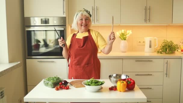 エネルギッシュな高齢者コーカサス女性が手を挙げると 料理しながらキッチンで踊り 新鮮でカラフルなサラダを準備することによって喜びが広がります 高品質のフルHd映像 — ストック動画