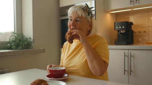 キッチンのテーブルで朝食にチョコレートでクロワッサンを食べる魅力的なエネルギッシュな高齢者カフカシアン60S女性 朝の儀式を楽しむアロマカプチーノコーヒーを飲む女性 — ストック動画