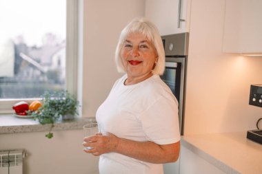 Gülümseyen güzel kadın mutfak masasına yaslanmış bir bardak su tutuyor. Yüksek kalite fotoğraf