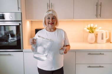 Gülümseyen son sınıf öğrencisi kadın filtrelenmiş suyu mutfağın iç kısımlarına döküyor. Bir bardak tatlı su tutan, günlük kıyafetli, çekici yaşlı kadın, fotokopi odası. Yüksek kalite fotoğraf