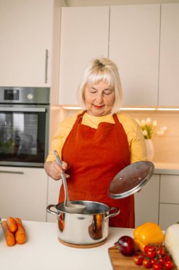 Güzel mutlu son sınıf güzeli mutfakta yemek pişiriyor ve ocağın tavasındaki çorbayı test ediyor. Yüksek kalite fotoğraf