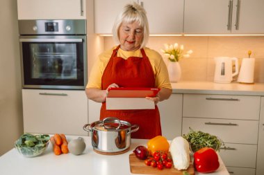 60 'lı yaşlardaki enerjik beyaz kadın mutfakta tablet bilgisayar kullanıyor ve taze sebzelerle yemek pişiriyor.