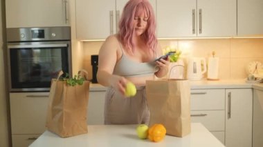 Şişman kadın çevrimiçi ev yemeği dağıtıyor. İnternetten yemek sipariş ediyorum. Beyaz mutfak iç kısımlarındaki ahşap masa üzerinde organik gıdalar bulunan el işi kağıt torbası. 