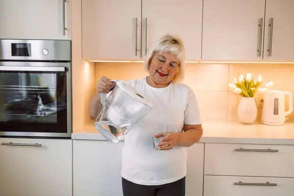 Smiling senior woman pouring filtered water into glass, kitchen interior. Attractive older woman in casual outfit holding glass of fresh water, copy space. High quality photo