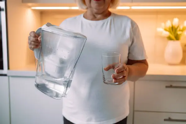 Smiling senior woman pouring filtered water into glass, kitchen interior. Attractive older woman in casual outfit holding glass of fresh water, copy space. High quality photo