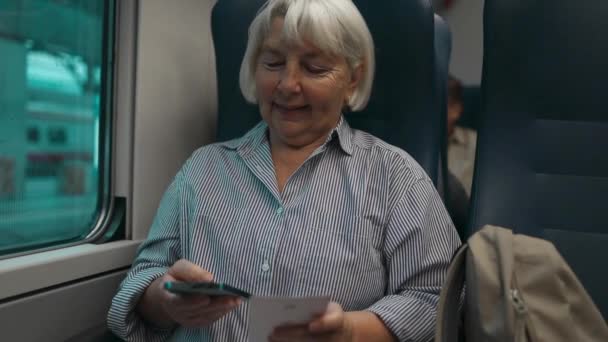 60代のシニアブロンドの髪白人女性が列車の窓に座って 彼女の旅の間に携帯電話を使ってチケットを持っています — ストック動画