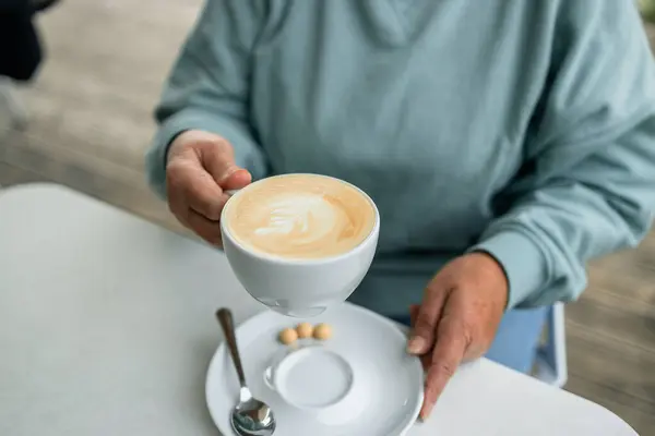 Woman holding cup of coffee with foam over wooden table sitting in city cafe. High quality photo