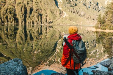 Dağın tepesinde dikilip Morskie Oko Gölü 'ne bakan 50' li yaşlarda kırmızı ceketli bir kadının arka plan çekimi. Sırt çantalı bir kadın dağ yamacında ve Polonya 'nın güzel manzarasının tadını çıkarıyor, Tatry. 