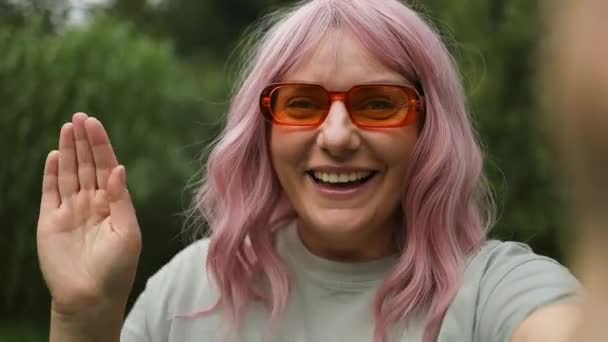 ブログのためのビデオを記録するオレンジのサングラスの美しい若いピンクの髪の女性を笑顔のコーカサス 女性が裏庭でウェブカムを通じてビデオ通話や手を振る — ストック動画