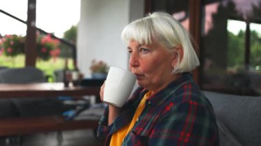 Yalnız, düşünceli, 60 'lı yaşlarda beyaz bir kadın arka bahçedeki kanepede, yumuşak renkli yastıklar ve çiçek saksılarıyla çay içiyor. Yaz bahçesinde güzel güneşli bir akşam. Yüksek kalite