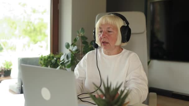 魅力的なインフルエンサー60代女性の肖像画 ヘッドフォンとラップトップを使用して笑顔 学習言語 オンラインウェビナー オーディオコース Eラーニング教育 — ストック動画