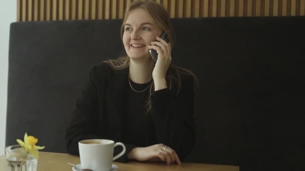 年轻快乐的女商人30多岁穿着西服 拿着手机聊天 拿着杯子喝茶 一个人坐在咖啡店餐厅的桌子旁 自由职业办公室业务的概念 — 图库视频影像