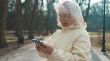 Elinde telefon olan neşeli, yaşlı, aktif bir kadın açık havada eğlenmeyi seviyor. Parktaki bankta otururken akıllı telefona bakan bir kadın. Modern teknolojiyi kullanarak. Yüksek kalite