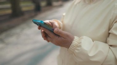 Neşeli yaşlı, yaşlı, aktif kadın elleri elinde telefonla dışarıda eğlenmeyi seviyor. Parktaki bankta otururken akıllı telefona bakan bir kadın. Modern teknolojiyi kullanarak. Yüksek