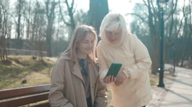 Parktaki bankta oturmuş akıllı telefonlara bakarken, iki yaşlı, aktif ve arkadaş canlısı kadın birbirleriyle konuşuyor. Parkta cep telefonu kullanarak yürüyen arkadaşlar.
