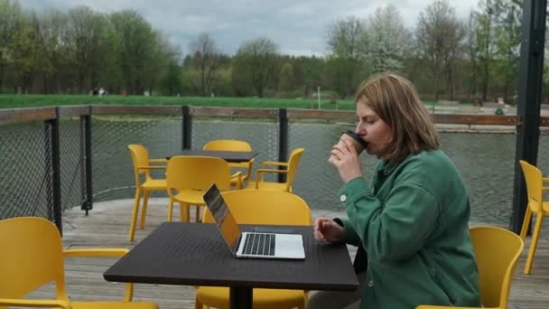 一个面带微笑的20多岁的女人正在享受一杯咖啡 她用笔记本电脑在时尚的城市咖啡店里做远程工作 笑漂亮的女孩喝热茶 靠近点 — 图库视频影像
