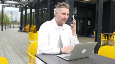 Mutlu Avrupalı işadamı akıllı telefondan bir telefon görüşmesi yapıyor, dışarıda şehir caddesindeki kafede oturup konuşuyor ve gülümsüyor. Yüksek kaliteli FullHD görüntüler