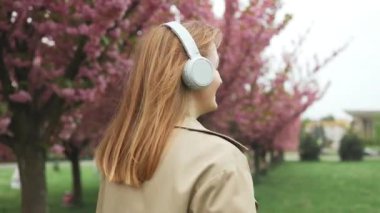 Neşeli, beyaz, sarışın, 20 'li yaşlarda gezgin bir kadın dışarıda kablosuz kulaklıklarla müzik dinleyerek dans ediyor. Mutlu gülümseyen kız modern kulaklıklarda eğleniyor. Yüksek kalite FullHD