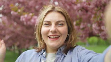Güzel, mutlu 30 'lu yaşlarda bir kadın çiçek açan sakura ağaçlarıyla parkta selfie çekiyor. Japon çiçek kavramının bahar seyahatleri,