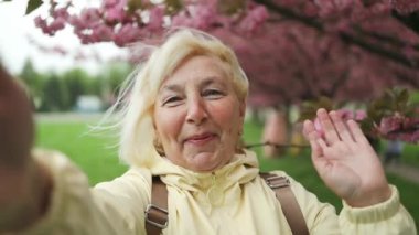 Güzel mutlu gezgin 50 'li yaşlardaki yaşlı kadın çiçek açan sakura ağaçlarıyla parkta selfie çekiyor. Japon çiçek kavramının bahar seyahatleri,
