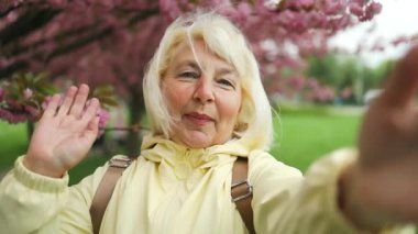 Güzel mutlu gezgin 50 'li yaşlardaki yaşlı kadın çiçek açan sakura ağaçlarıyla parkta selfie çekiyor. Japon çiçek kavramının bahar seyahatleri,
