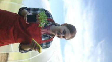 Kadın çiftçi dijital telefonla domates bitkilerini kontrol ediyor. Çiftçi arka bahçedeki seranın yanında domates bitkileriyle akıllı telefon kullanıyor. Modern teknoloji uygulaması tarımsal büyüme faaliyetlerinde. Yüksek