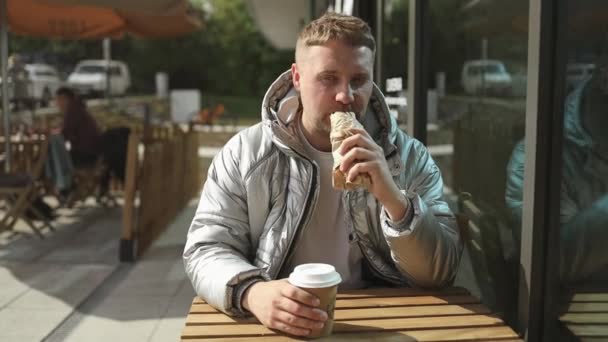 一个英俊的白人年轻人坐在户外咖啡馆里吃着美味的热狗的画像 送货员 食物可口 优质Fullhd影片 — 图库视频影像
