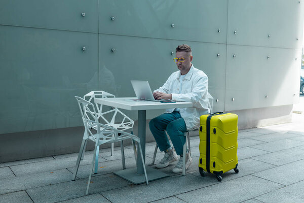 Happy traveling Caucasian man in stylish clothes uses mobile smartphone and laptop, checking trip destination on Internet. Male waiting at flight gates for plane boarding, smiling have a vacation trip