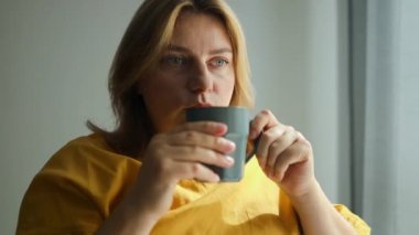 Evde kahve içerken gülümseyen genç bir kadının portresi. İş kadınları sabah kahvelerini içerken iş yerinde oturur ve sabahın erken saatlerinde güneşin doğuşunun tadını çıkarırlar. Yüksek kalite FullHD